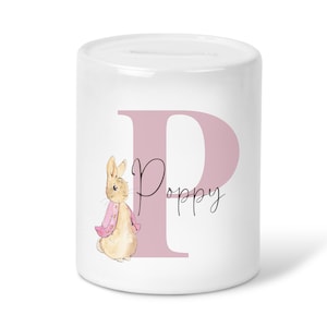 May include: White ceramic money box with a pink letter P and the name Poppy written in cursive. A cartoon bunny wearing a pink jacket is to the left of the letter.