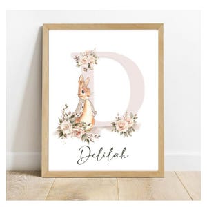 Pode incluir: Uma impressão emoldurada com uma ilustração em aquarela de um coelho em pé na frente de uma letra D rosa com detalhes florais. O nome "Delilah" está escrito abaixo da letra.