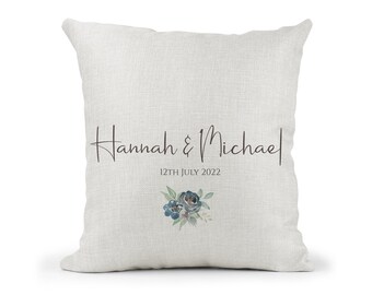 May include: A white linen pillow with the text "Hannah & Michael" and "12th July 2022" in black script. A small blue watercolor floral design is below the text.