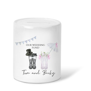 May include: White ceramic money box with a wedding theme. It features a bride and groom wearing boots, a white umbrella, and the text "Our Wedding Fund" and "Tom and Becky".