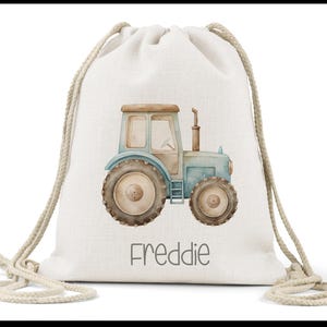May include: A cream-coloured drawstring backpack made of a woven fabric, featuring a watercolour illustration of a blue and brown tractor in the centre. Below the tractor, the name Freddie is printed in a simple, dark grey sans-serif font.