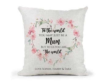 May include: A square, off-white throw pillow with a floral heart design. The heart is composed of pink flowers and green leaves. Text reads "To the world you may just be a Mum but to us you are the world." The names "LOVE SOPHIE, HARRY & TARA" are at the bottom.