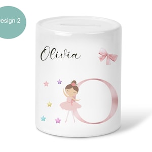 May include: White ceramic money box with a ballerina design. The box features the name "Olivia" in script, a pink bow, and a watercolor ballerina with a pink tutu. Colourful stars and a large pink letter "O" complete the design.