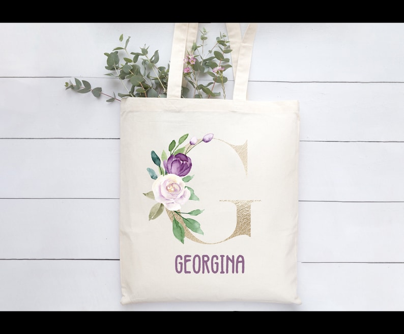 May include: Cream-colored tote bag featuring a gold letter "G" and the name "GEORGINA" in purple. The bag is adorned with a watercolor floral design in purple, white, and green, with eucalyptus and pink flowers in the background.