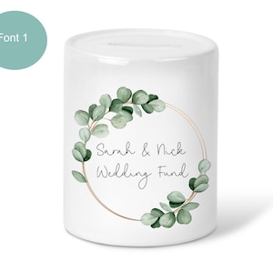 May include: White ceramic money box with a gold circle and green eucalyptus leaves around the edge. The text "Sarah & Nick Wedding Fund" is written in black cursive font.
