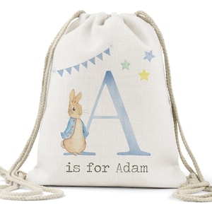 May include: A white canvas drawstring bag with a blue letter A, a Peter Rabbit illustration, and the text "is for Adam". The bag has a blue and white bunting and yellow stars.
