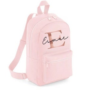 May include: A pale pink backpack with a front pocket and a top handle. The backpack features a rose gold letter "E" and the name "Esmae" in a cursive font. The bag has adjustable shoulder straps and a zippered main compartment.