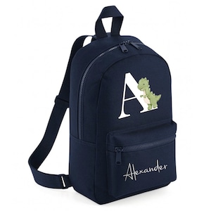 May include: A navy blue backpack with a front zip pocket and adjustable straps. It features a white letter 'A' with a green dinosaur illustration. The name 'Alexander' is written in cursive below the pocket.