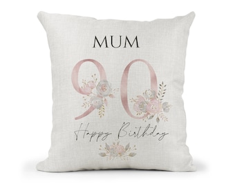 May include: A square, off-white linen pillow with the word "MUM" in black at the top. The number "90" is in a dusty rose color, decorated with floral accents. The words "Happy Birthday" are written in a script font.