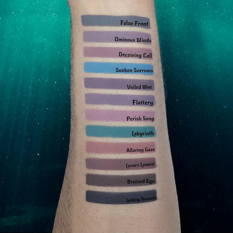 May include: A series of lipstick swatches in various shades of purple, blue, and pink are displayed on an arm. Each swatch has a name written in white text, including "False Front," "Ominous Winds," and "Lurking Beneath."