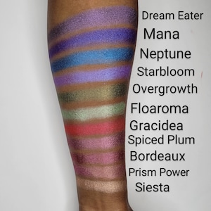 Puede incluir: Un primer plano de un brazo con 12 muestras de sombra de ojos diferentes. Las muestras est&aacute;n etiquetadas de arriba a abajo: Dream Eater, Mana, Neptune, Starbloom, Overgrowth, Floaroma, Gracidea, Spiced Plum, Bordeaux, Prism Power, Siesta.