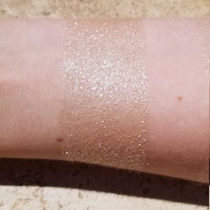May include: A swatch of shimmering, golden-bronze makeup applied to the inner forearm. The product has a glittery, metallic finish, creating a luminous effect. The skin tone is fair, and the product is applied in a rectangular shape.