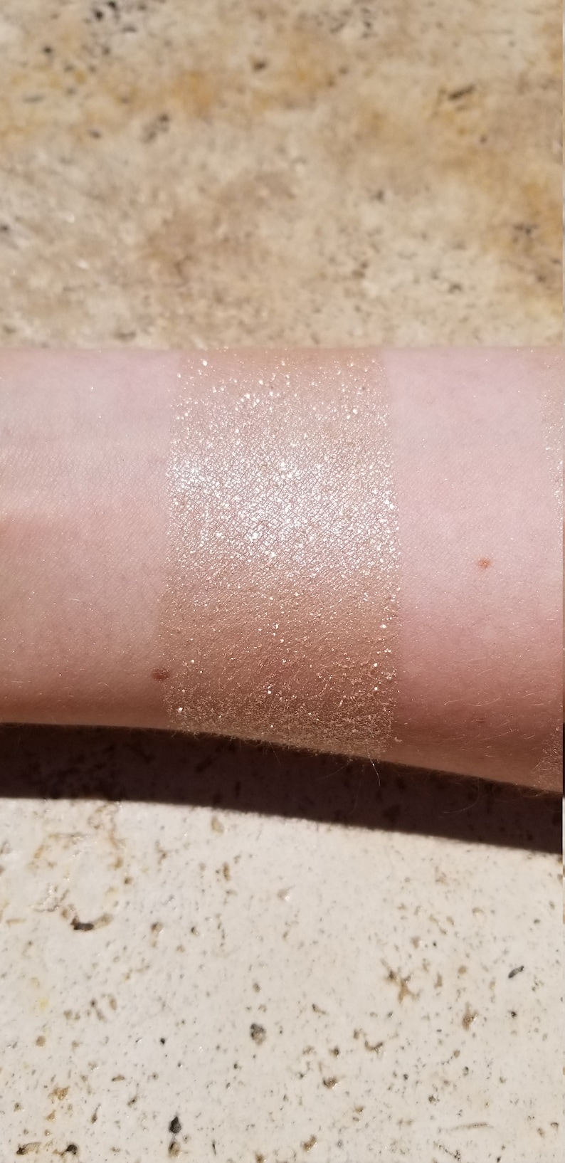 May include: A swatch of shimmering, golden-bronze makeup applied to the inner forearm. The product has a glittery, metallic finish, creating a luminous effect. The skin tone is fair, and the product is applied in a rectangular shape.