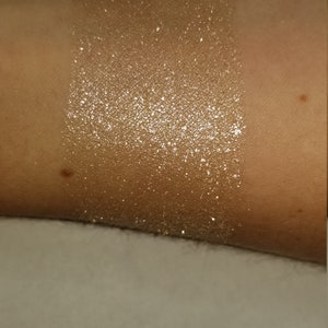 May include: A swatch of shimmering, golden-toned glitter applied to skin. The glitter is densely packed, creating a bright, reflective surface. The skin tone is a warm, medium shade. The glitter is applied in a rectangular shape.