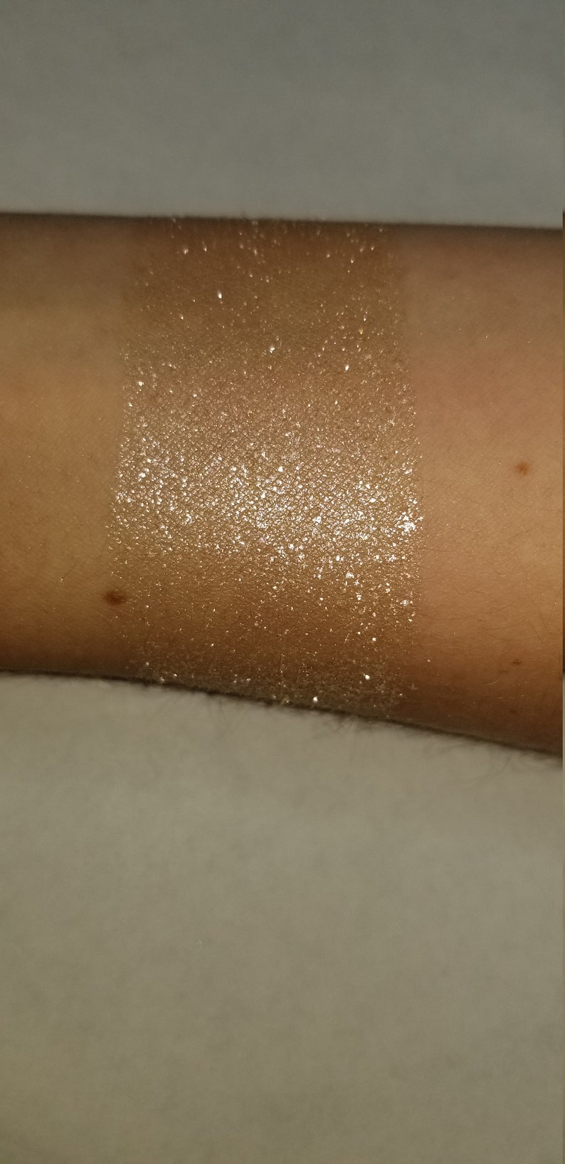 May include: A swatch of shimmering, golden-toned glitter applied to skin. The glitter is densely packed, creating a bright, reflective surface. The skin tone is a warm, medium shade. The glitter is applied in a rectangular shape.