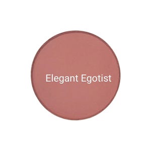 Puede incluir: Un compacto de rubor redondo y rosa con el texto "Elegant Egotist" impreso en la superficie.