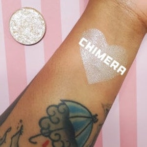 May include: A close-up of a silver eyeshadow compact next to an arm with a heart-shaped swatch of shimmering eyeshadow. The word "CHIMERA" is printed on the arm. The arm has a tattoo of a blue umbrella with red raindrops.