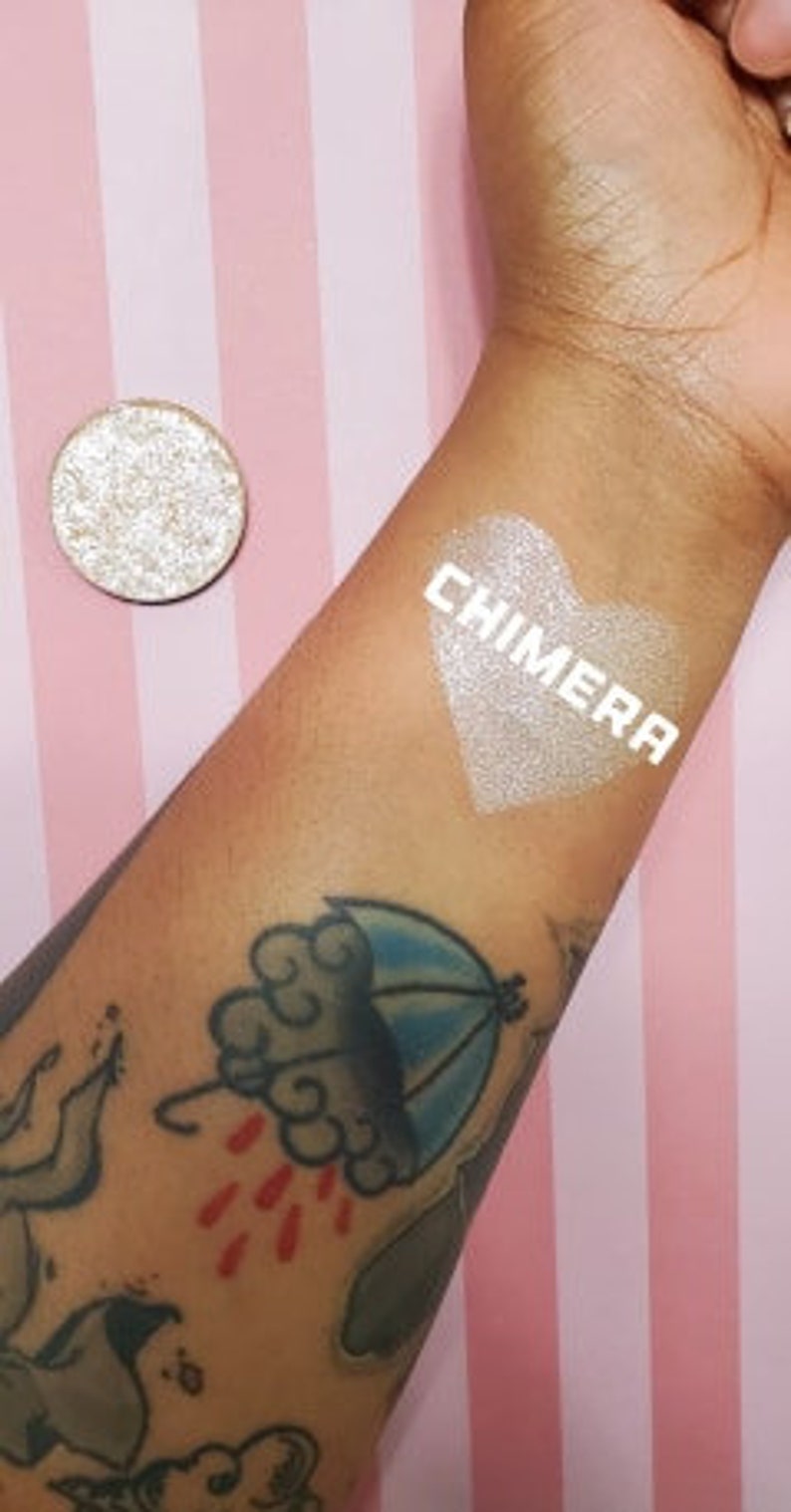 May include: A close-up of a silver eyeshadow compact next to an arm with a heart-shaped swatch of shimmering eyeshadow. The word "CHIMERA" is printed on the arm. The arm has a tattoo of a blue umbrella with red raindrops.