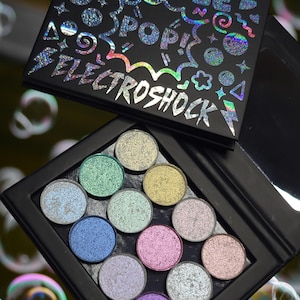 May include: A black eyeshadow palette with the text "BUBBLE POP! ELECTROSHOCK" in holographic lettering. The palette contains 12 circular eyeshadow pans in various shimmering colours, including green, blue, and pink. The palette is open, revealing the eyeshadows.