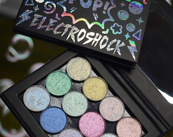 May include: A black eyeshadow palette with the text "BUBBLE POP! ELECTROSHOCK" in holographic lettering. The palette contains 12 circular eyeshadow pans in various shimmering colours, including green, blue, and pink. The palette is open, revealing the eyeshadows.