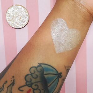 May include: A round, shimmering eyeshadow pan next to a heart-shaped swatch on an arm. The eyeshadow and swatch are a light, metallic color. The arm has tattoos, including a cloud and umbrella design. The background is pink and white striped.