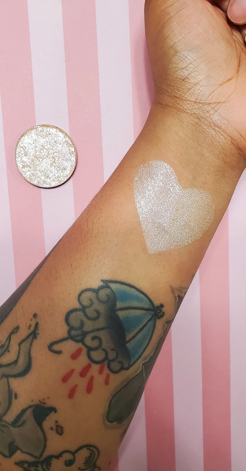 May include: A round, shimmering eyeshadow pan next to a heart-shaped swatch on an arm. The eyeshadow and swatch are a light, metallic color. The arm has tattoos, including a cloud and umbrella design. The background is pink and white striped.