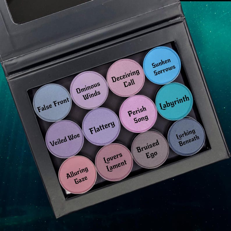 May include: A black eyeshadow palette with twelve circular pans. Each pan is a different color, ranging from blue to pink, and labeled with a unique name like "False Front" and "Labyrinth". The palette is open, revealing the colorful shadows.