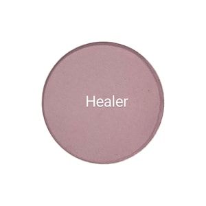 Puede incluir: Un producto de maquillaje redondo de color malva con la palabra "Healer" impresa en blanco. El producto tiene un acabado mate y se presenta sobre un fondo blanco.
