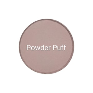 May include: A round, light brown powder compact with the words "Powder Puff" in white text. The compact has a slightly raised edge and a matte finish. The background is plain white.