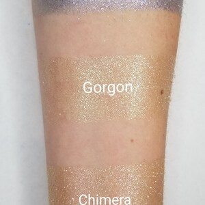 May include: Three shimmering eyeshadow swatches on an arm. The top swatch is a sparkling lavender color labeled "Scylla." The middle swatch is a gold color labeled "Gorgon." The bottom swatch is a light gold color labeled "Chimera."