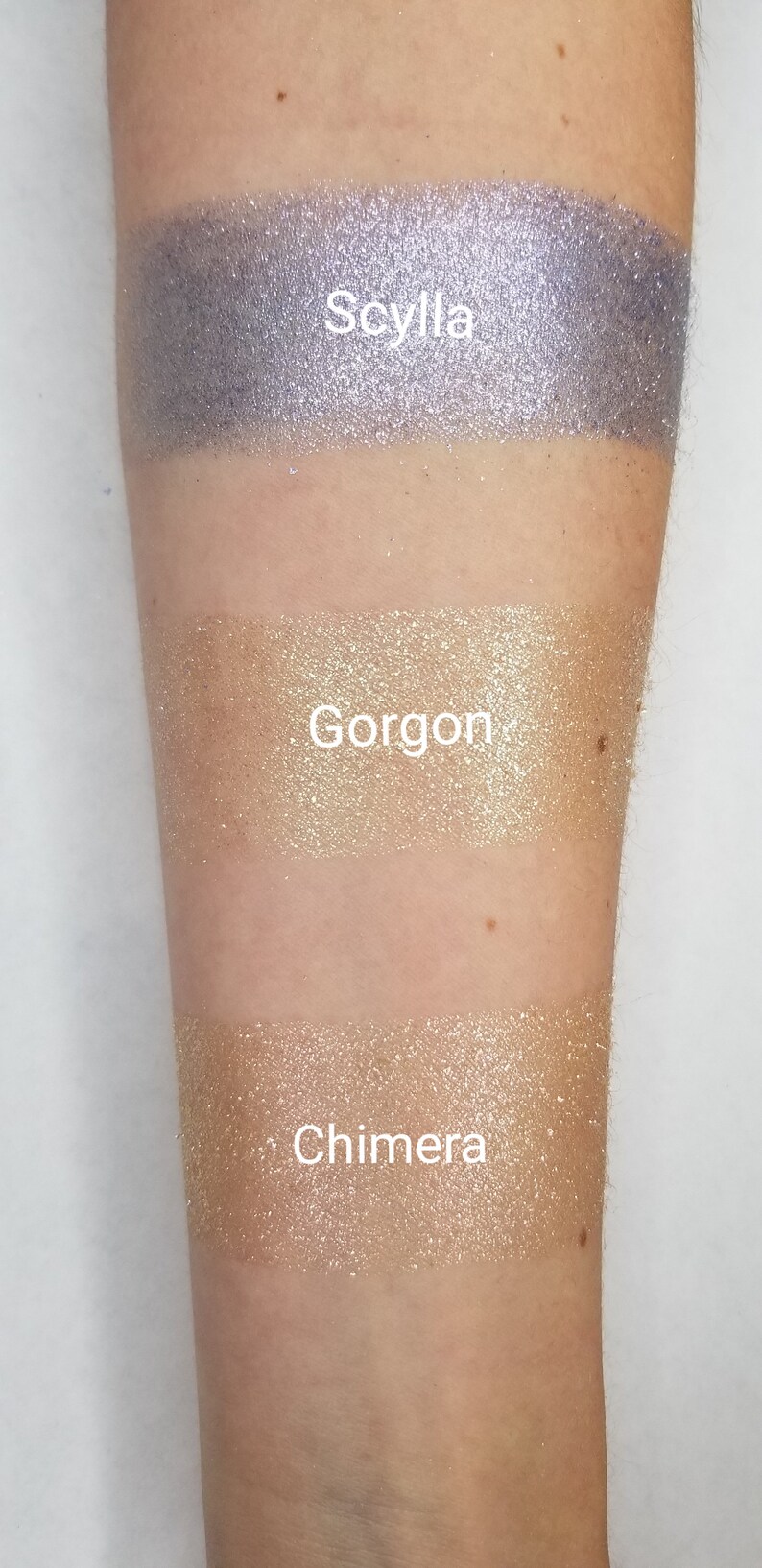 May include: Three shimmering eyeshadow swatches on an arm. The top swatch is a sparkling lavender color labeled "Scylla." The middle swatch is a gold color labeled "Gorgon." The bottom swatch is a light gold color labeled "Chimera."