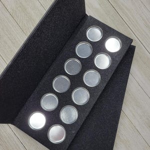 Pode incluir: Uma paleta de maquilhagem preta, com textura de glitter, com 12 recipientes circulares prateados. A paleta está aberta, revelando os recipientes vazios inseridos numa inserção de espuma preta. A paleta está angulada numa superfície de madeira clara.
