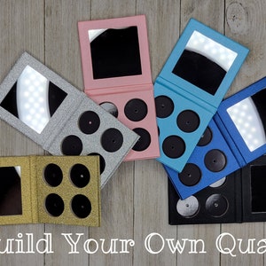 Puede incluir: Cinco paletas de maquillaje magnéticas con espejos y bandejas extraíbles. Las paletas son de color plateado, dorado, rosa, azul y negro. El texto "Build Your Own Quad" está en la parte inferior de la imagen.
