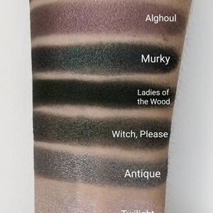Op de afbeelding: Een close-up van iemands arm met swatches van oogschaduw in verschillende tinten groen, grijs en bruin. De swatches zijn gelabeld met de namen "Carrie", "Alghoul", "Murky", "Ladies of the Wood", "Witch, Please", "Antique" en "Twilight".