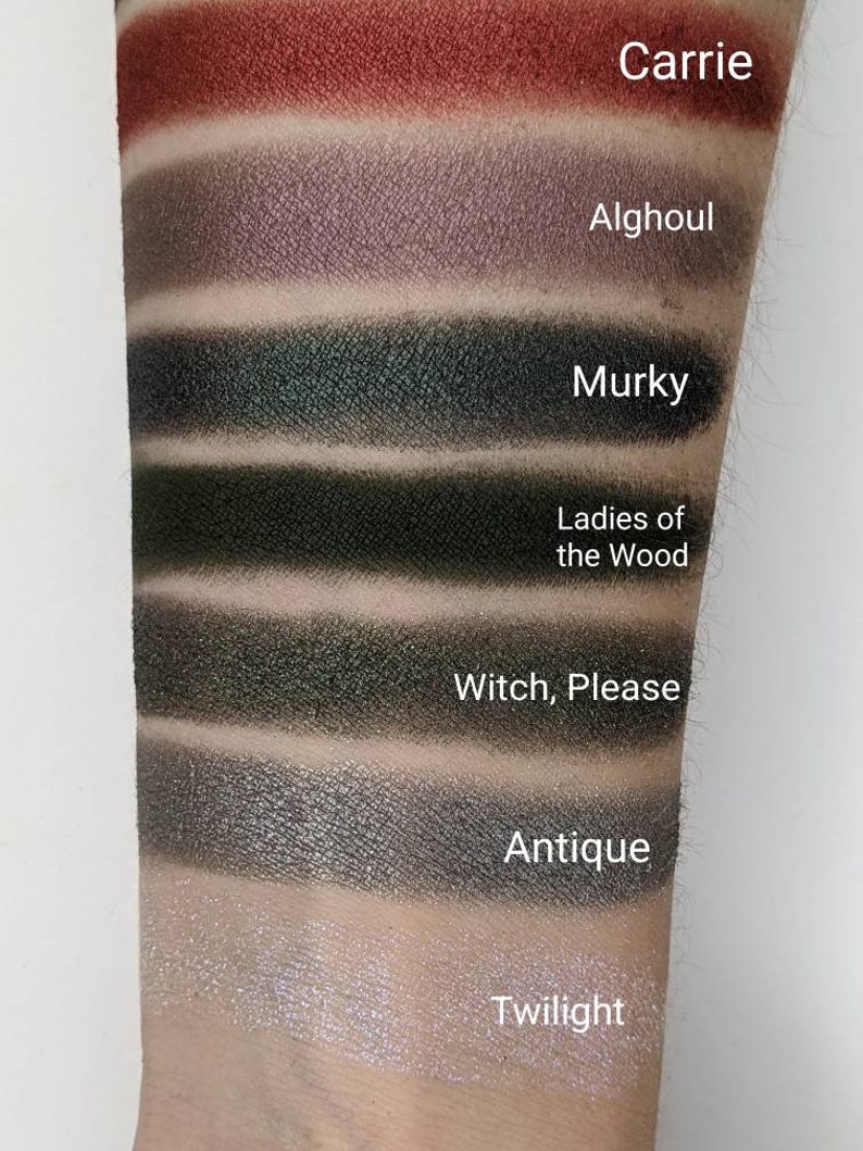 Op de afbeelding: Een close-up van iemands arm met swatches van oogschaduw in verschillende tinten groen, grijs en bruin. De swatches zijn gelabeld met de namen "Carrie", "Alghoul", "Murky", "Ladies of the Wood", "Witch, Please", "Antique" en "Twilight".