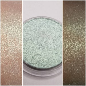 May include: A close-up of three swatches of eyeshadow. The center swatch is a light green, shimmery eyeshadow in a clear plastic container. The other two swatches are of a similar eyeshadow in different shades of gold.