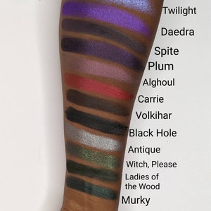 Op de afbeelding: Een close-up van iemands arm met swatches van oogschaduw in verschillende tinten paars, rood, zwart, groen en blauw. De swatches zijn gelabeld met namen zoals "Twilight", "Daedra", "Spite", "Plum", "Alghoul", "Carrie", "Volkihar", "Black Hole", "Antique", "Witch, Please", "Ladies of the Wood" en "Murky".