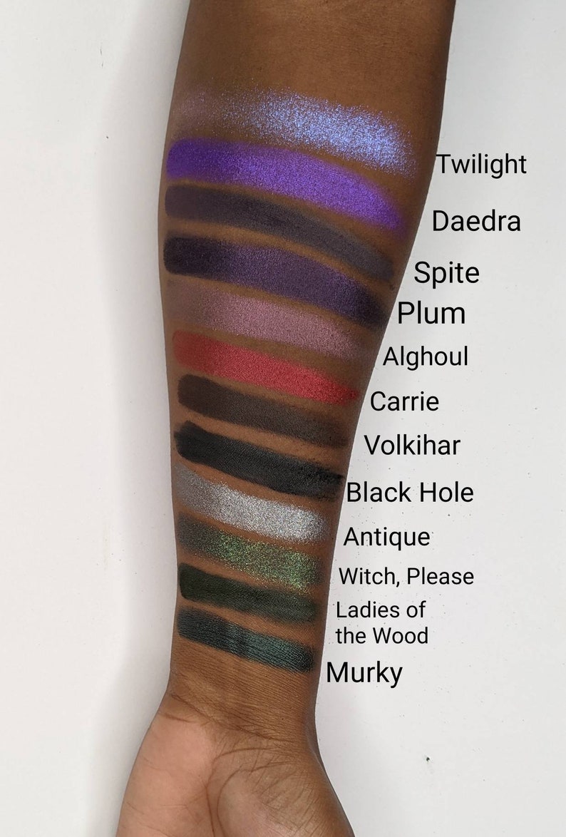 Op de afbeelding: Een close-up van iemands arm met swatches van oogschaduw in verschillende tinten paars, rood, zwart, groen en blauw. De swatches zijn gelabeld met namen zoals "Twilight", "Daedra", "Spite", "Plum", "Alghoul", "Carrie", "Volkihar", "Black Hole", "Antique", "Witch, Please", "Ladies of the Wood" en "Murky".