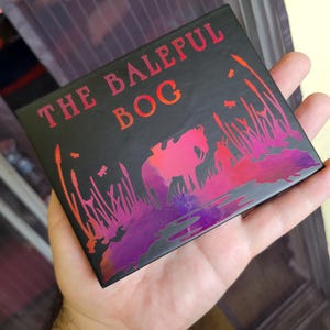 Op de afbeelding: Een zwarte doos met een holografische afbeelding van een paard in een moeras. De doos heeft de tekst "The Baleful Bog" op de voorkant.