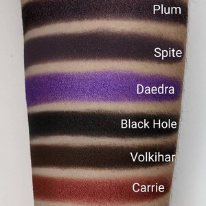 Op de afbeelding: Een close-up van een make-up palet met zeven oogschaduw swatches. De swatches zijn van boven naar beneden gelabeld: Plum, Spite, Daedra, Black Hole, Volkihar, Carrie en Alghoul.