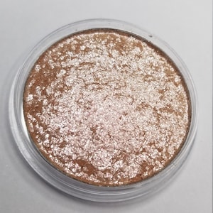 May include: A round, shimmering eyeshadow compact with a clear plastic lid. The eyeshadow has a textured, metallic appearance with shades of bronze and silver. The product is designed for eye makeup application.