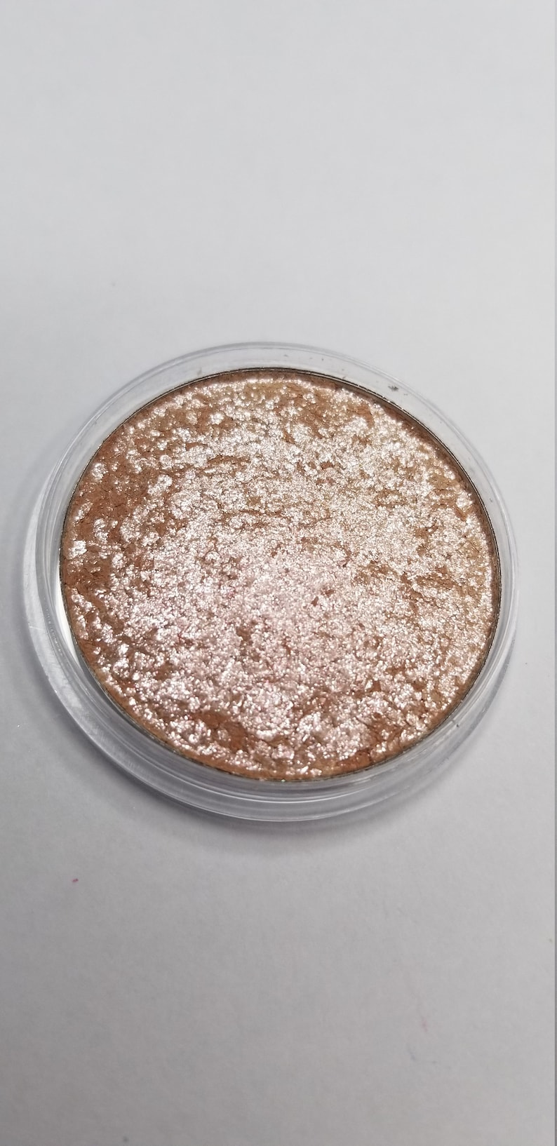 May include: A round, shimmering eyeshadow compact with a clear plastic lid. The eyeshadow has a textured, metallic appearance with shades of bronze and silver. The product is designed for eye makeup application.