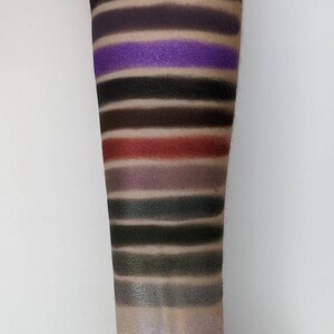 Op de afbeelding: Een close-up van iemands arm met swatches van oogschaduw in verschillende tinten zwart, bruin, paars, groen en zilver. De swatches zijn gerangschikt in rijen, met de donkerste tinten bovenaan en de lichtste tinten onderaan.