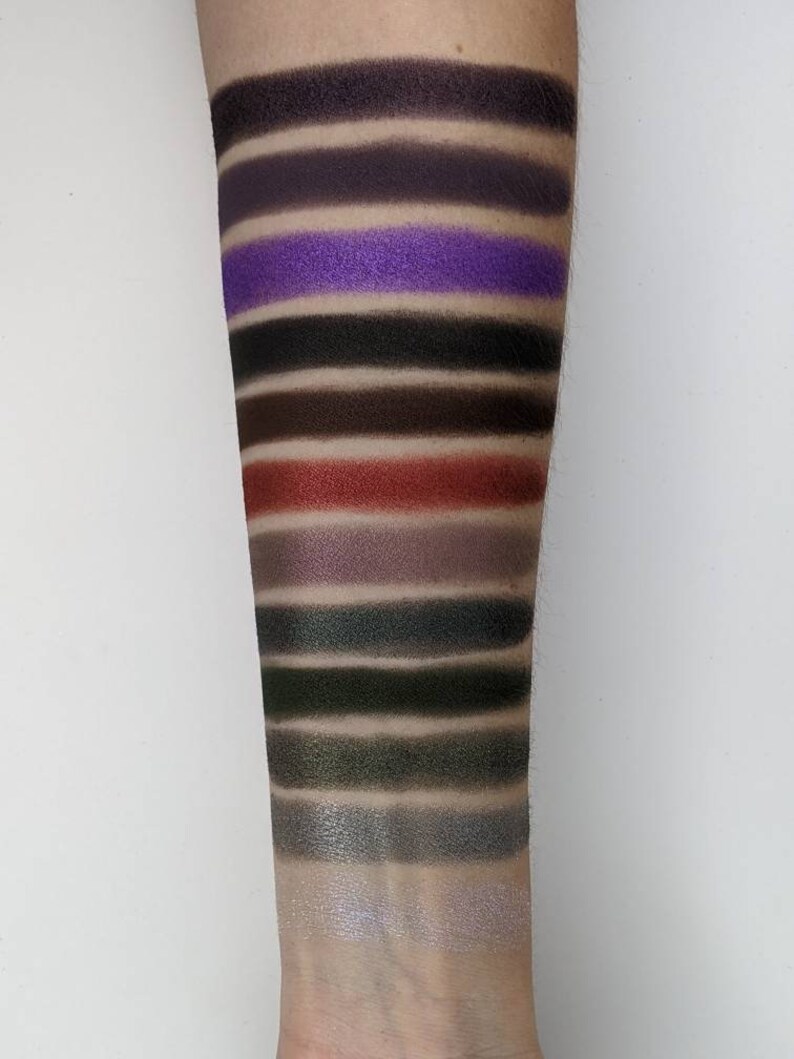 Op de afbeelding: Een close-up van iemands arm met swatches van oogschaduw in verschillende tinten zwart, bruin, paars, groen en zilver. De swatches zijn gerangschikt in rijen, met de donkerste tinten bovenaan en de lichtste tinten onderaan.