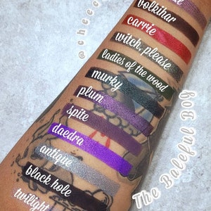 Op de afbeelding: Een close-up van een arm met swatches van oogschaduw in verschillende tinten paars, groen, rood en zwart. De swatches zijn gelabeld met namen zoals "Alghoul", "Volkihar", "Carrie", "Witch, Please", "Ladies of the Wood", "Murky", "Plum", "Spite", "Daedra", "Antique", "Black Hole" en "Twilight". De tekst "The Baleful Bog" staat op de arm.