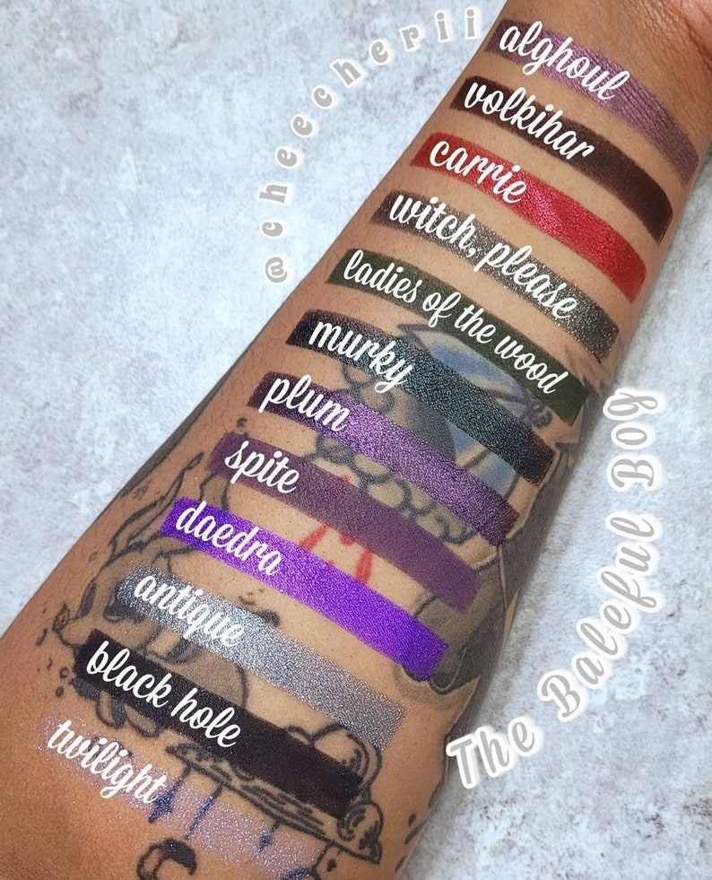 Op de afbeelding: Een close-up van een arm met swatches van oogschaduw in verschillende tinten paars, groen, rood en zwart. De swatches zijn gelabeld met namen zoals "Alghoul", "Volkihar", "Carrie", "Witch, Please", "Ladies of the Wood", "Murky", "Plum", "Spite", "Daedra", "Antique", "Black Hole" en "Twilight". De tekst "The Baleful Bog" staat op de arm.