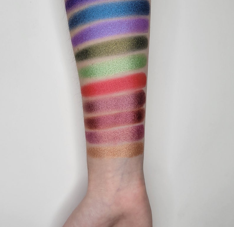 Puede incluir: Un primer plano de un brazo con muestras de sombra de ojos en varios colores, incluyendo azul, morado, verde, rojo, marr&oacute;n y dorado.