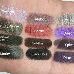Op de afbeelding: Een close-up van 12 oogschaduw swatches op een hand. De swatches zijn gelabeld met de namen "Twilight", "Alghoul", "Antique", "Witch, Please", "Carrie", "Daedra", "Ladies of the Wood", "Volkihar", "Spite", "Murky", "Black Hole" en "Plum".