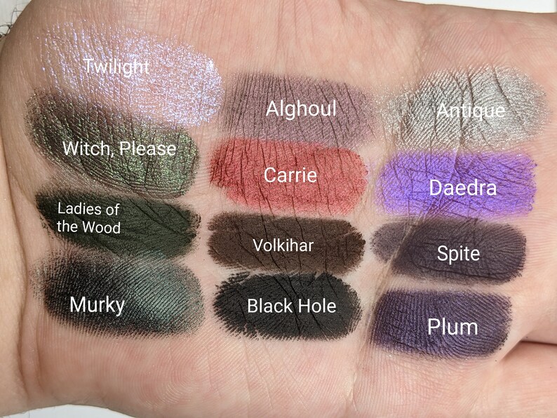 Op de afbeelding: Een close-up van 12 oogschaduw swatches op een hand. De swatches zijn gelabeld met de namen "Twilight", "Alghoul", "Antique", "Witch, Please", "Carrie", "Daedra", "Ladies of the Wood", "Volkihar", "Spite", "Murky", "Black Hole" en "Plum".