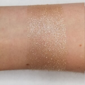 May include: A swatch of shimmering, golden-bronze eyeshadow applied to the inner forearm. The product has a high shimmer finish, reflecting light. The skin tone is fair, providing a contrast to the product.